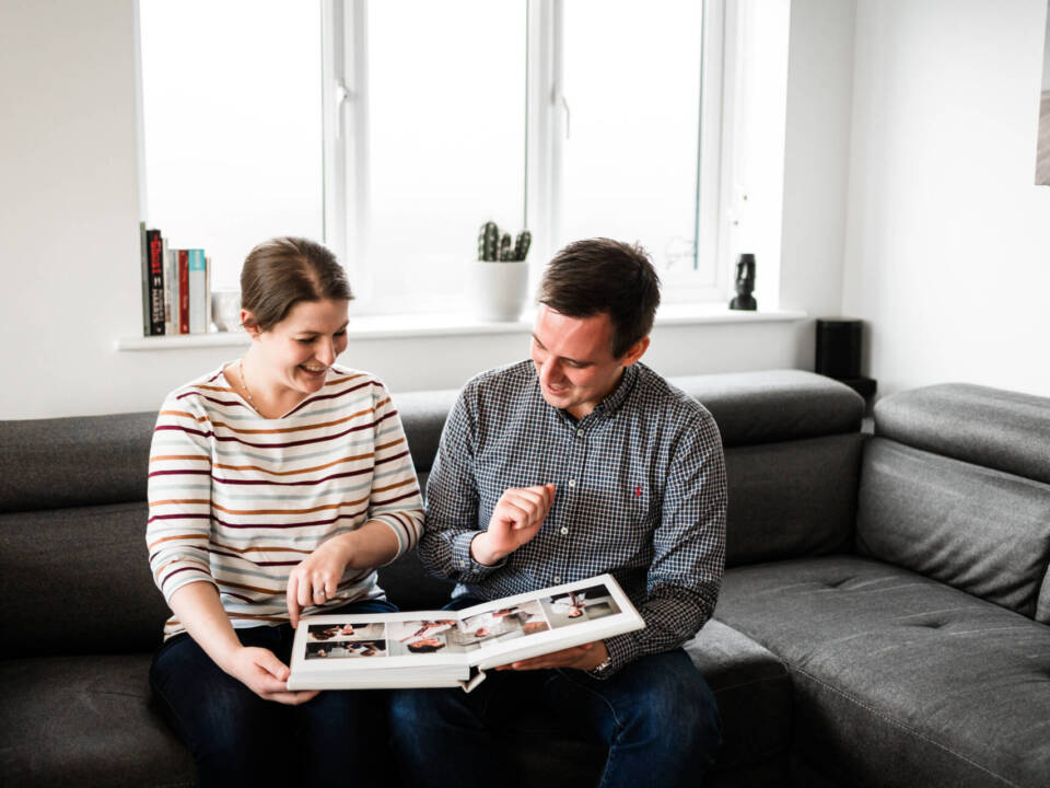 a man and woman sat on a sofa in a living room looking at a photo album