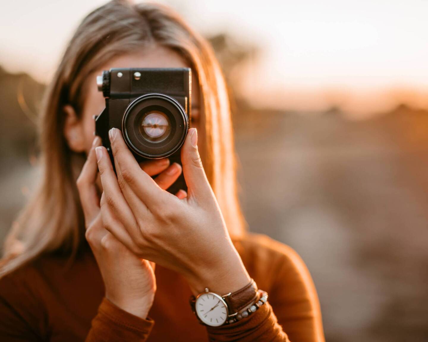 female photographer holding camera over face in front of sunset 