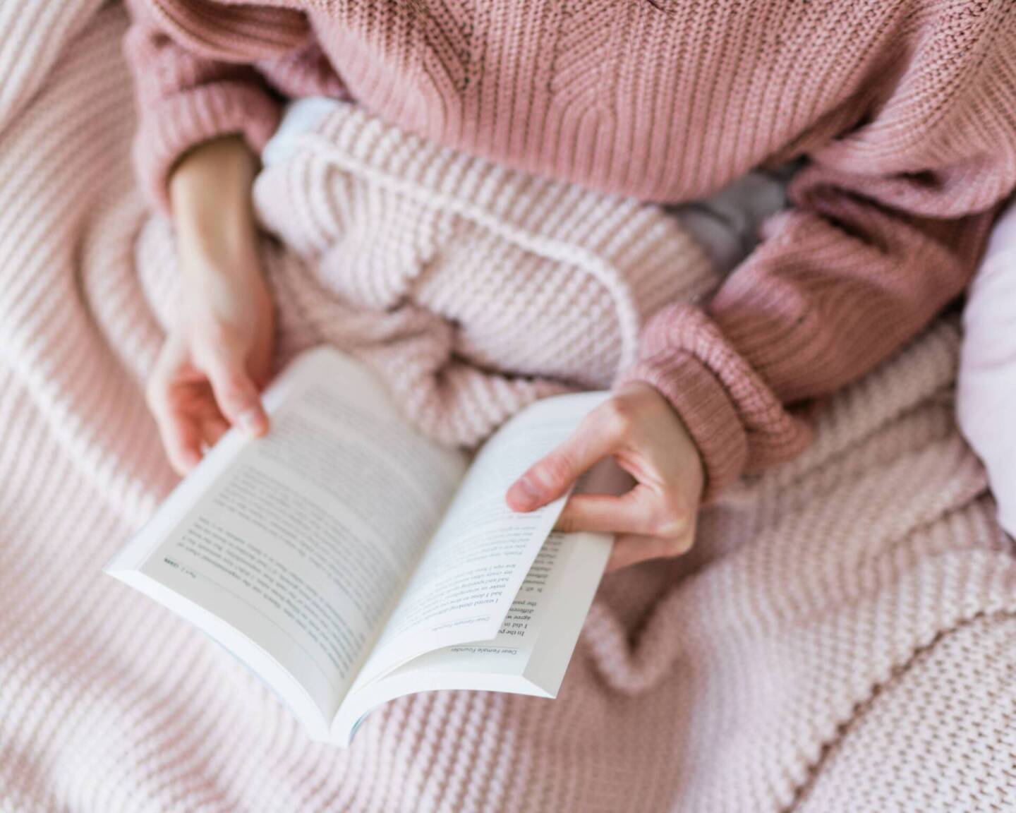 woman in pink jumper reading a book under a blanket 