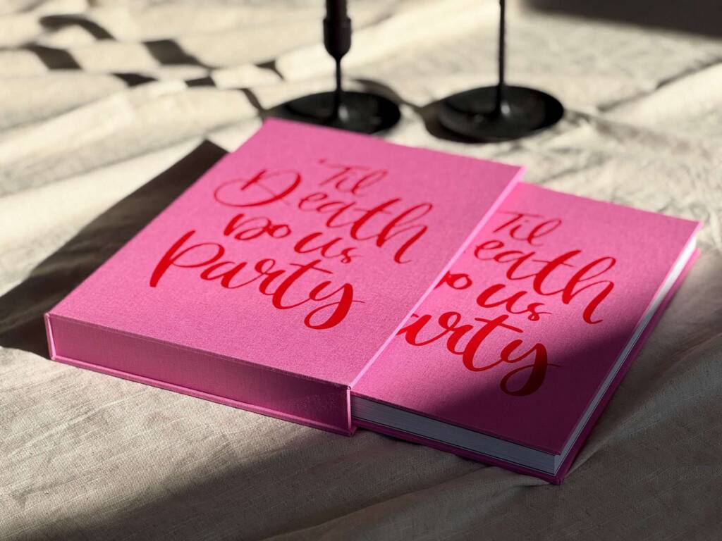 A pink Coffee Table Book and slipcase with red screenprinted cover design on a beige blanket with candle sticks in the background.